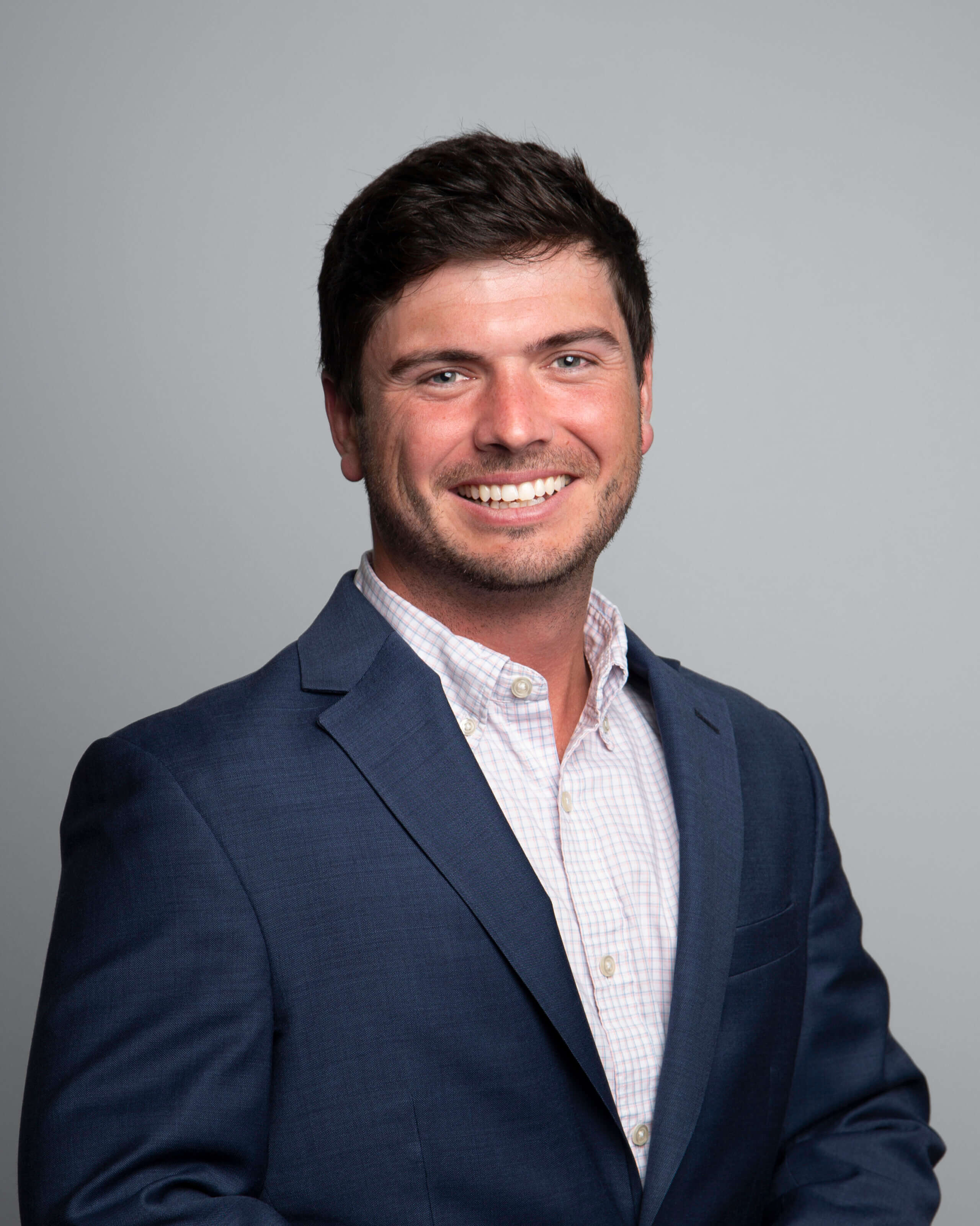 Christopher Dupré – RNGD leadership headshot of Christopher Dupré, wearing a navy suit and light checkered dress shirt, smiling confidently against a gray background.