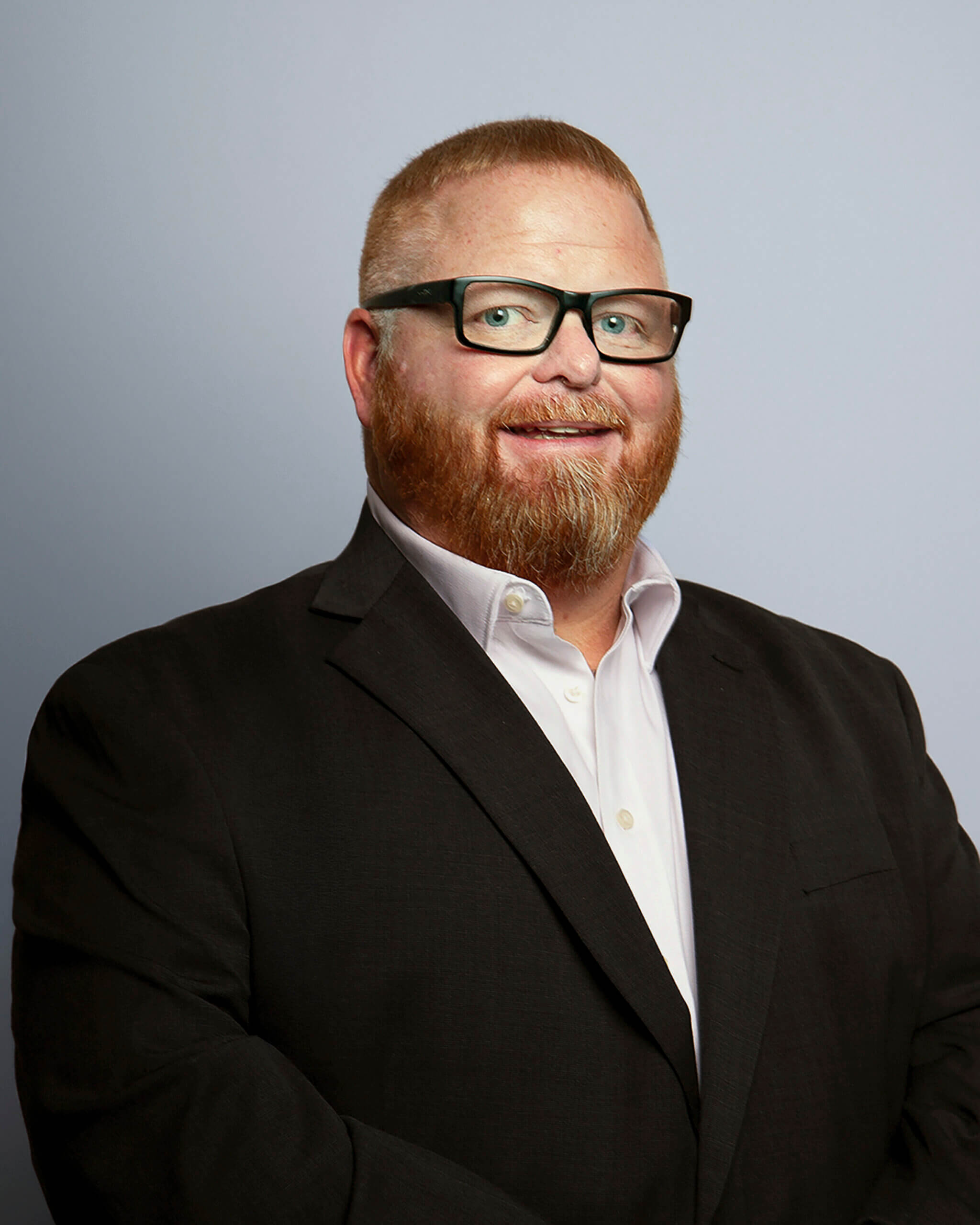 Jonathan Hill – Professional headshot of a man with short red hair and beard, wearing glasses, a black suit jacket, and a white dress shirt, smiling in front of a light gray background.
