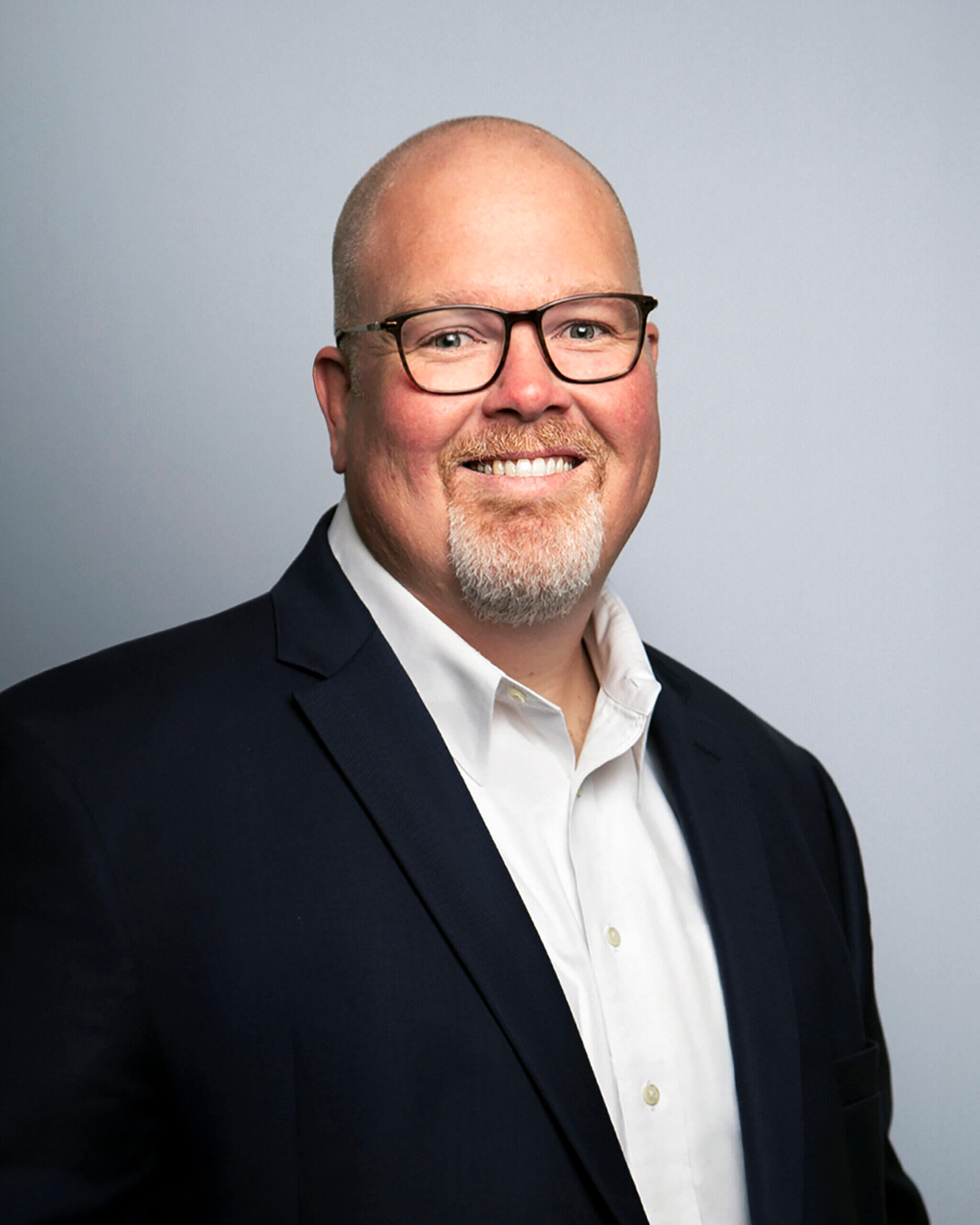 Josh Hill – RNGD executive headshot of Josh Hill, wearing glasses, a navy suit, and white shirt, smiling against a gray background.