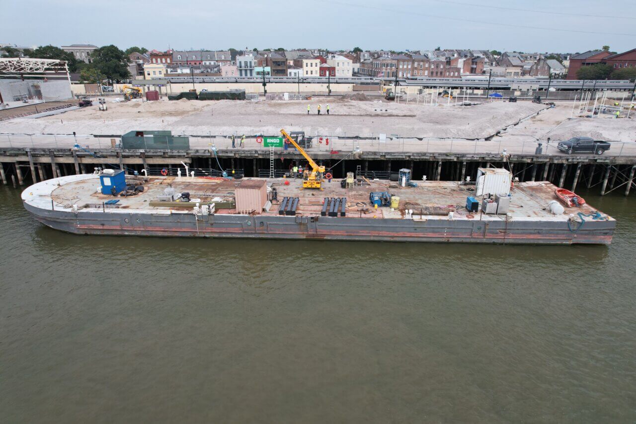 Construction barge positioned along Governor Nichols Wharf on the Mississippi River as crews repair damaged dock piles beneath the structure.