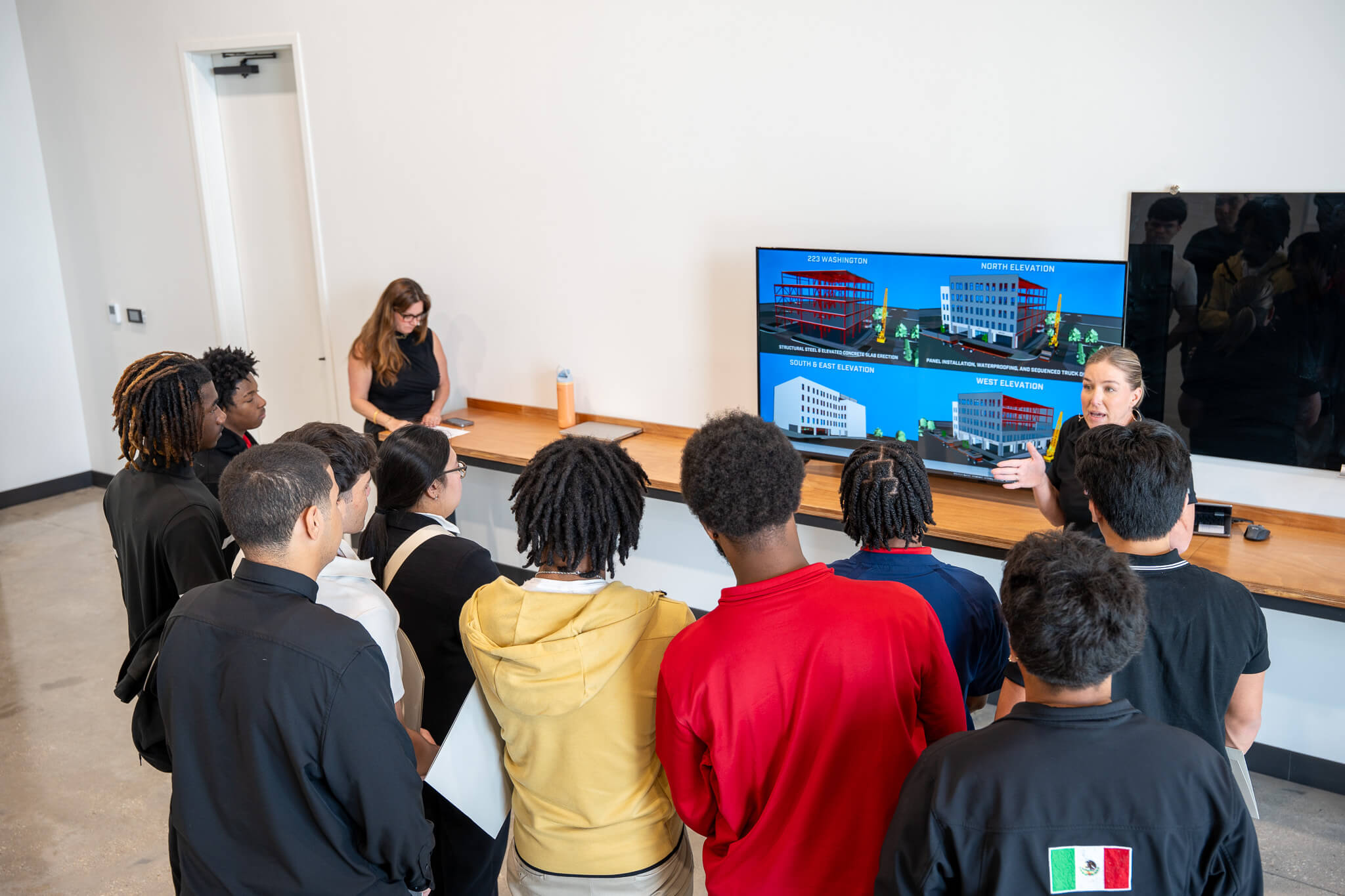 A group of students watches an RNGD team member present a digital construction model and project coordination visuals on a large display screen.