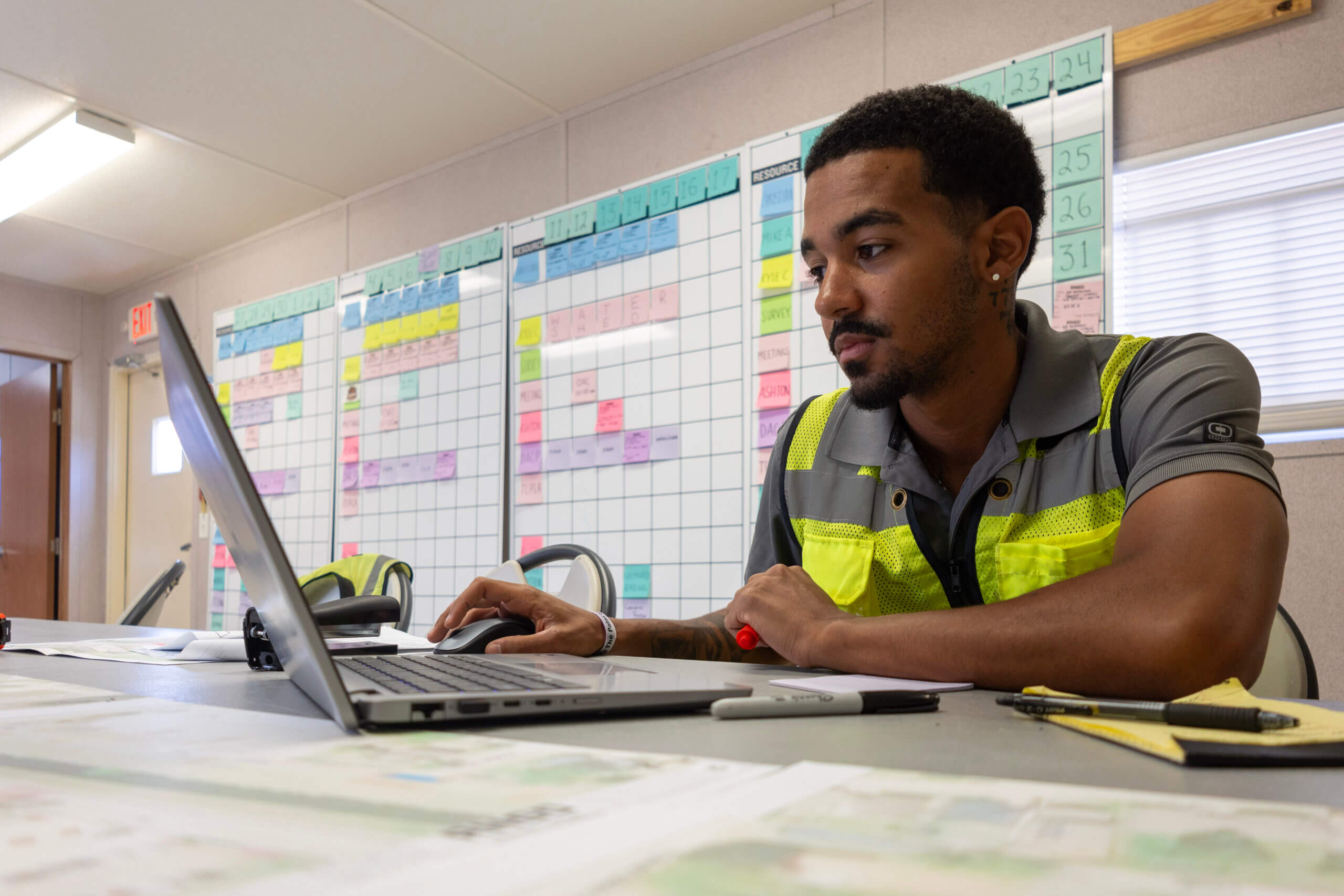 RNGD Project Engineer Isaiah Margan works on a laptop inside a field office trailer with construction schedules and planning boards displayed behind him.