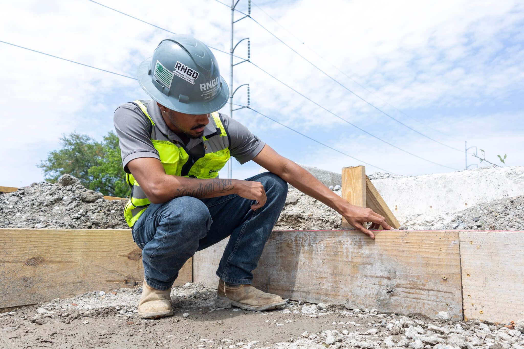 RNGD Project Engineer Isaiah Margan kneels at a roadway construction site inspecting formwork and layout lines while wearing a hard hat and high-visibility safety vest.