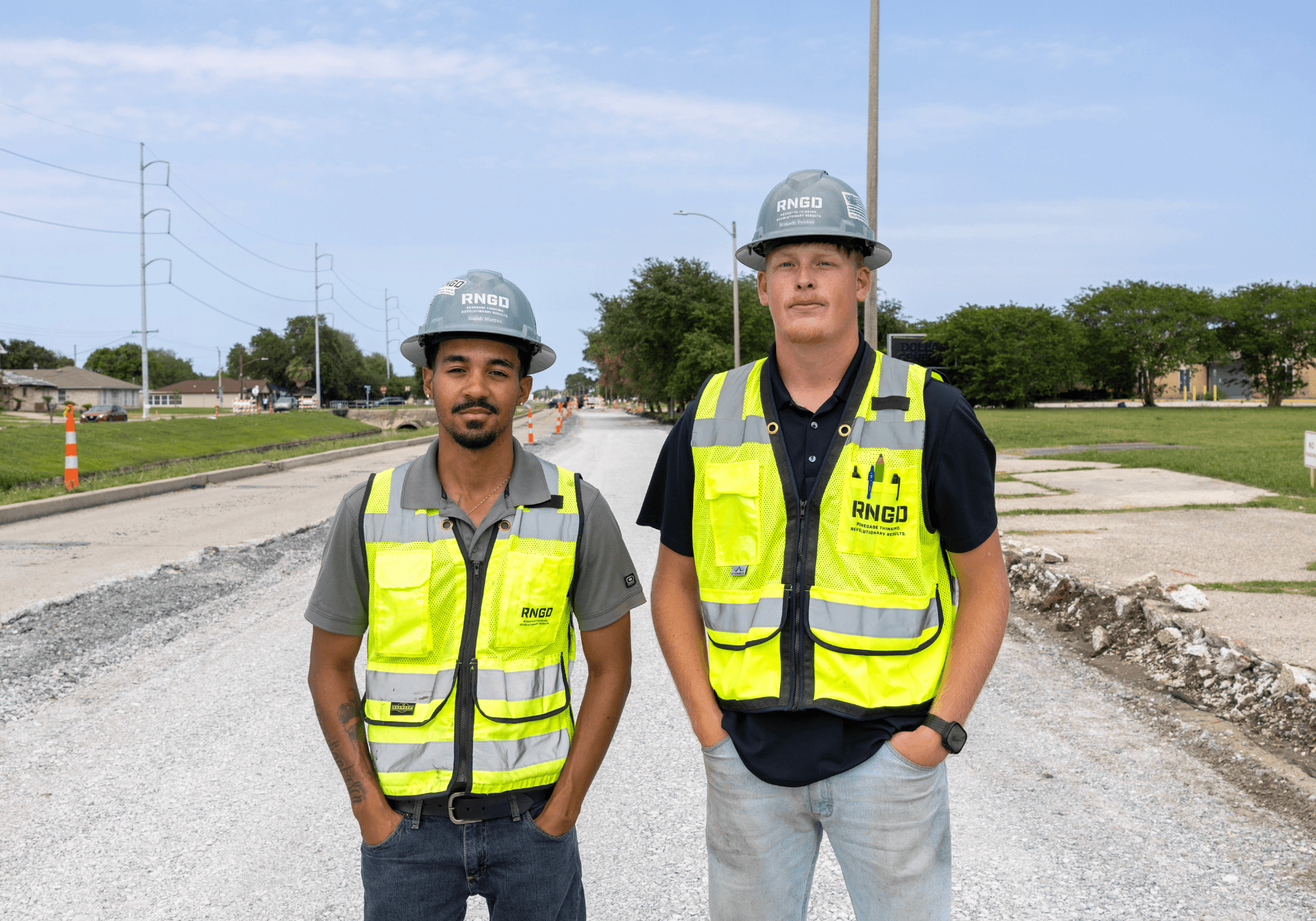 Two RNGD project engineers wearing hard hats and high-visibility safety vests stand on an active roadway construction project site, posing in the middle of a partially reconstructed street.